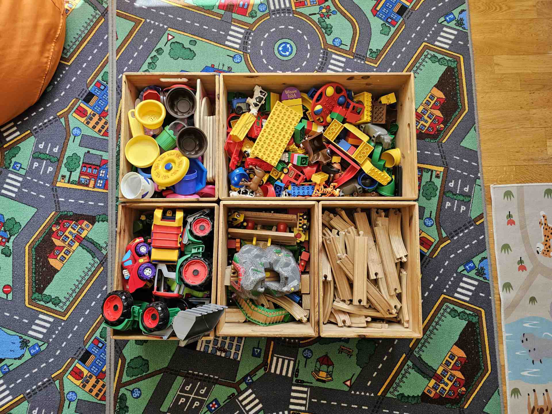Vier Holzkisten voller spannender Spielsachen, darunter Bauklötze, Spielzeugtraktoren und Holzeisenbahnen, sortiert auf einem bunten Straßenteppich.
