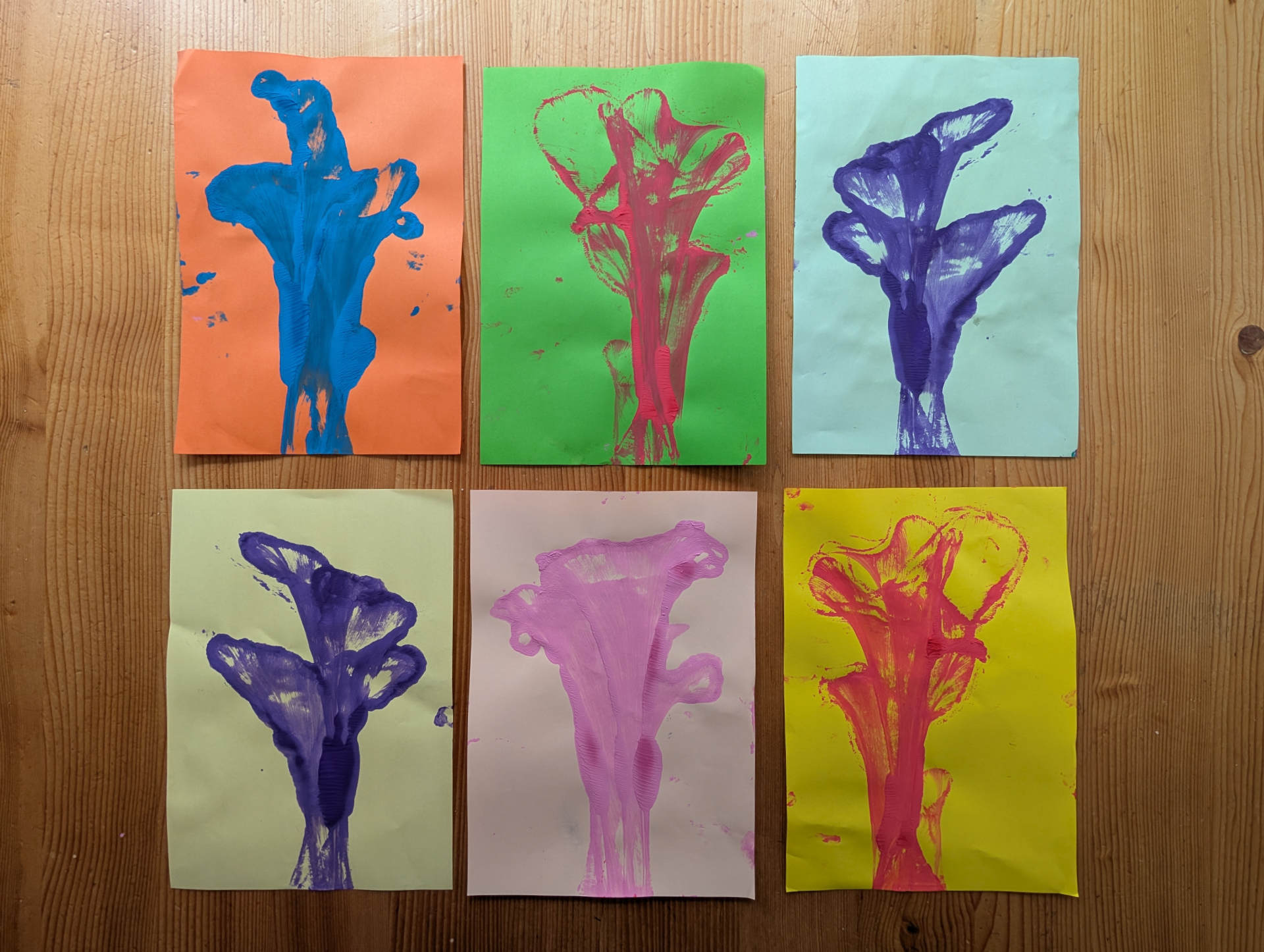 Sechs farbenfrohe, selbstgemalte Kunstwerke unserer Tageskinder, die bunte Blumenmotive auf verschiedenfarbigem Papier zeigen.