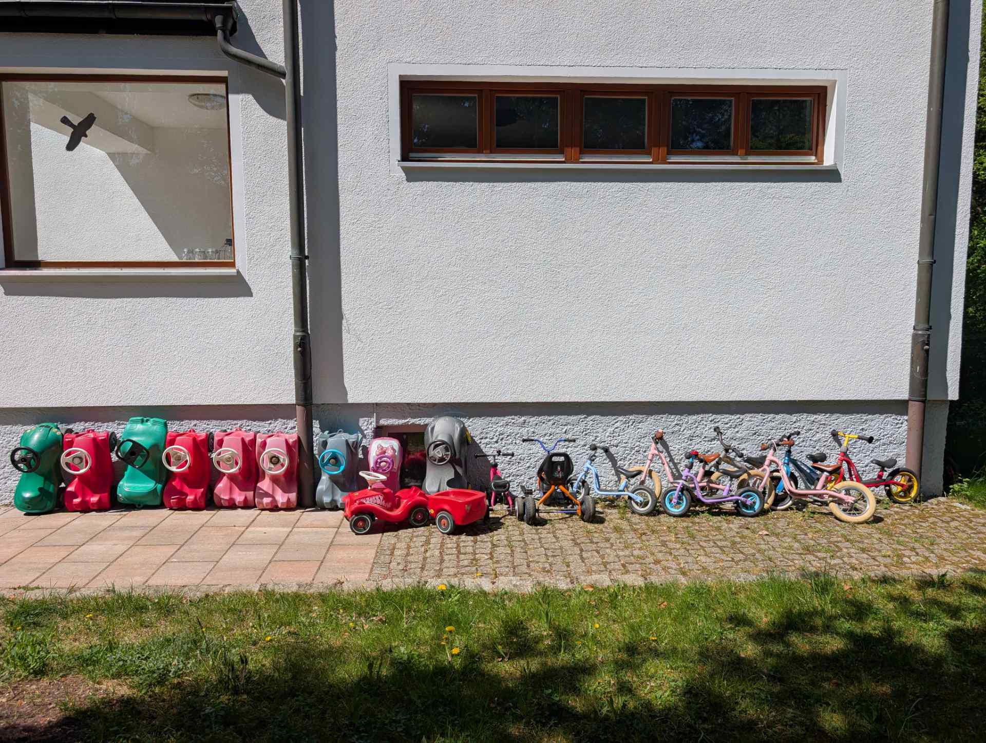 Eine lange Reihe bunter Bobbycars, Dreiräder und Laufräder parkt ordentlich vor dem Haus und wartet auf den nächsten Ausflug im Freien.