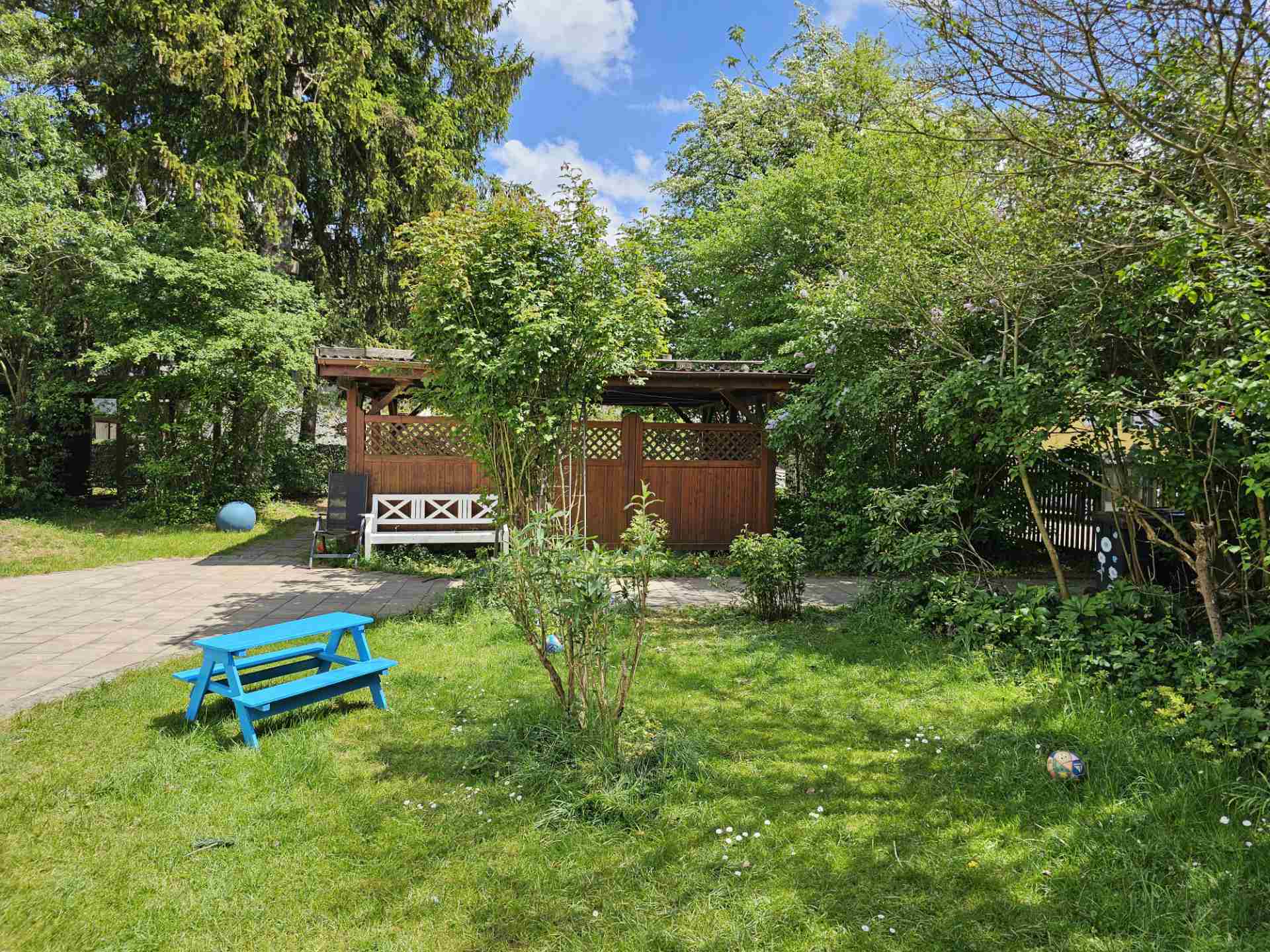 Weitläufiger Garten der Tagespflege mit viel grüner Rasenfläche zum Toben, einem überdachten Außenbereich und einer kleinen blauen Kinder-Sitzbank.