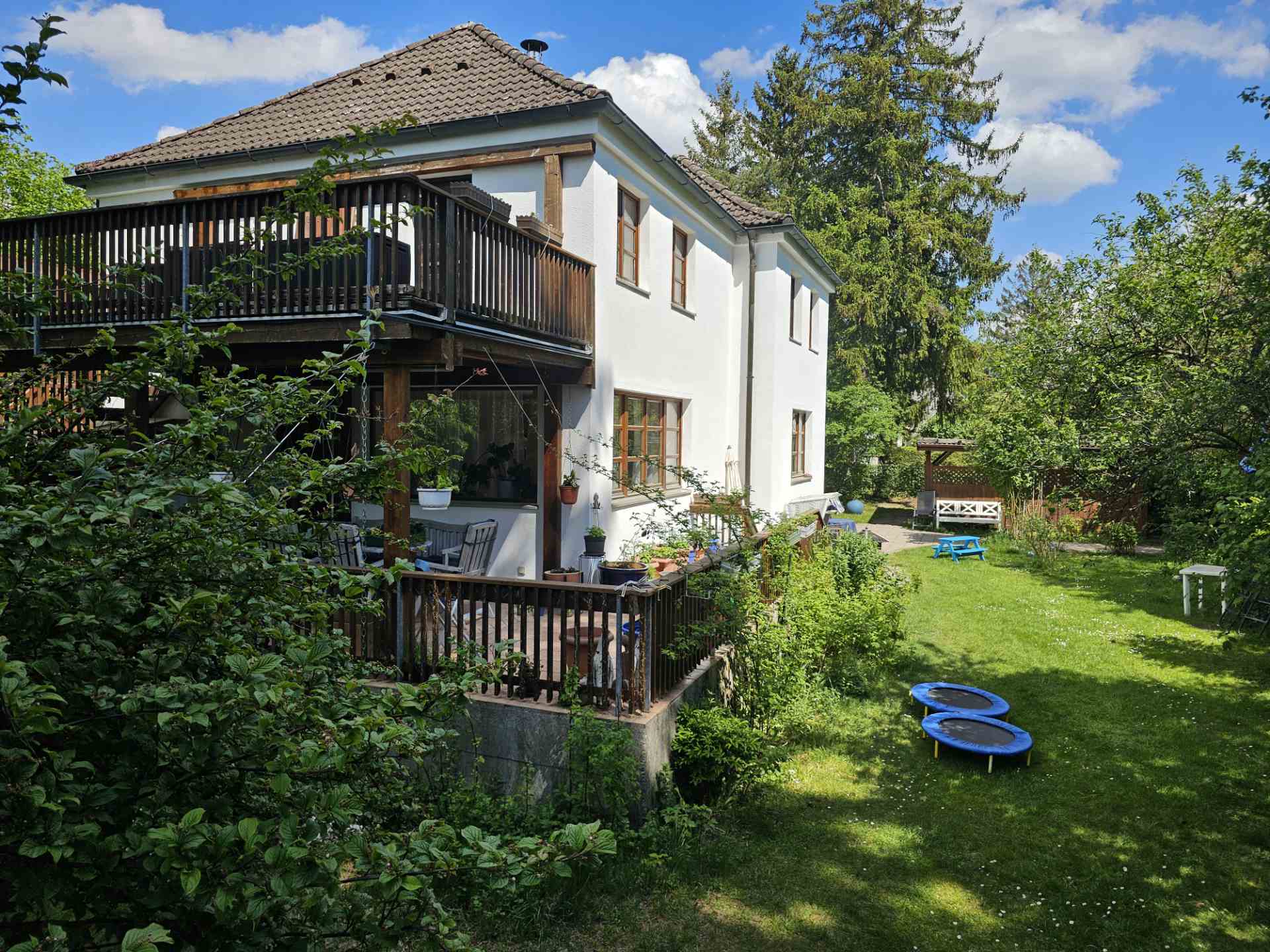 Seitenansicht unseres Hauses in Ottobrunn mit großem Holzbalkon und direktem Zugang in den großen Garten, in dem kleine Trampoline bereitstehen.